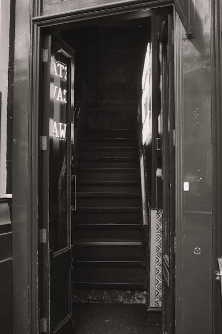 A narrow flight of wooden stairs leading upwards inside a building, viewed through an open doorway with black-painted door frames and hinges. The stairs are enclosed by a dark wooden wall on one side